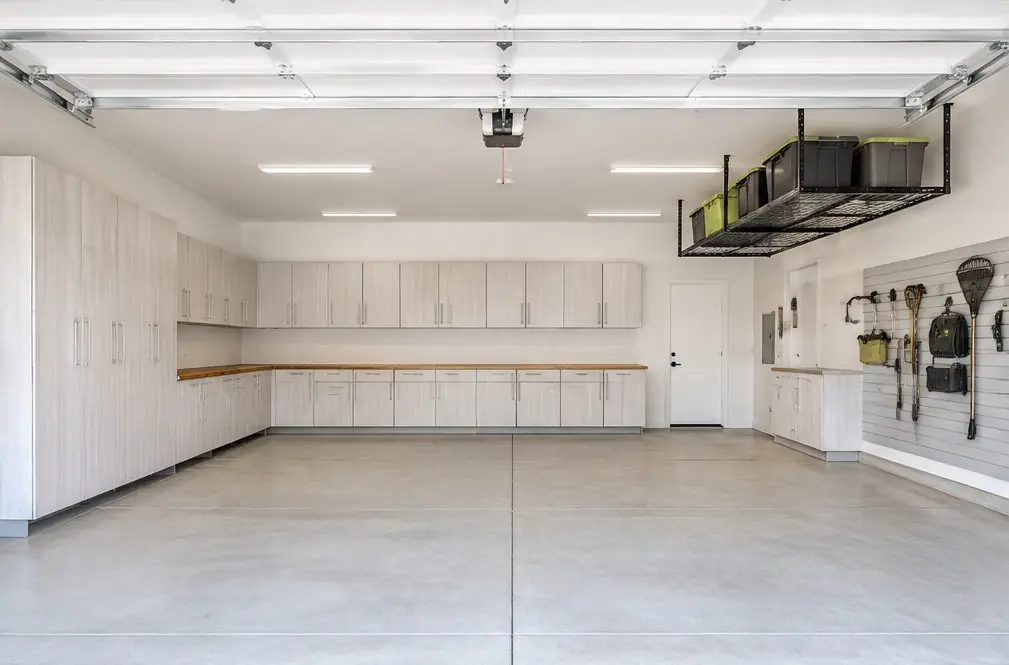 Illustration: a new three-car garage in a Menifee, CA tract home with light melamine tall, base and wall cabinets on the back and side walls, a long wood bench, a section of slatwall with hooks and a single ceiling rack over the third bay